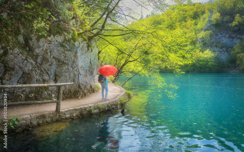 在春天的雨中，一个带着红色雨伞的女人在美丽的湖泊附近的小路上。克罗地亚普利特维斯湖。科罗拉多州