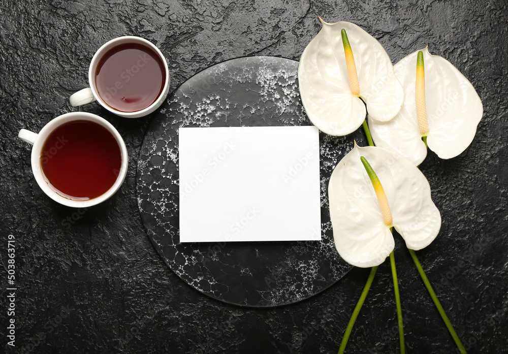 Beautiful anthurium flowers with cups of tea and sheet of paper on dark background