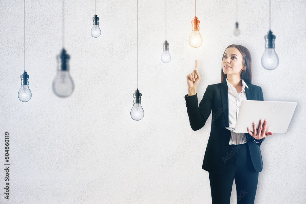 Attractive young european businesswoman with laptop pointing up while standing on concrete wall back