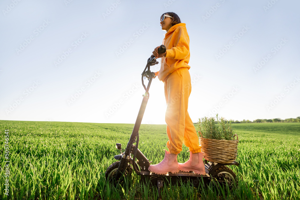 Young person riding electric scooter with plant in pot on green field during sunset. Concept of sust