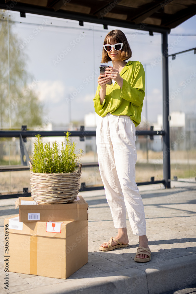 Young woman stands with parcels and flowerpot on a bus stop and using phone. Concept of sustainabili