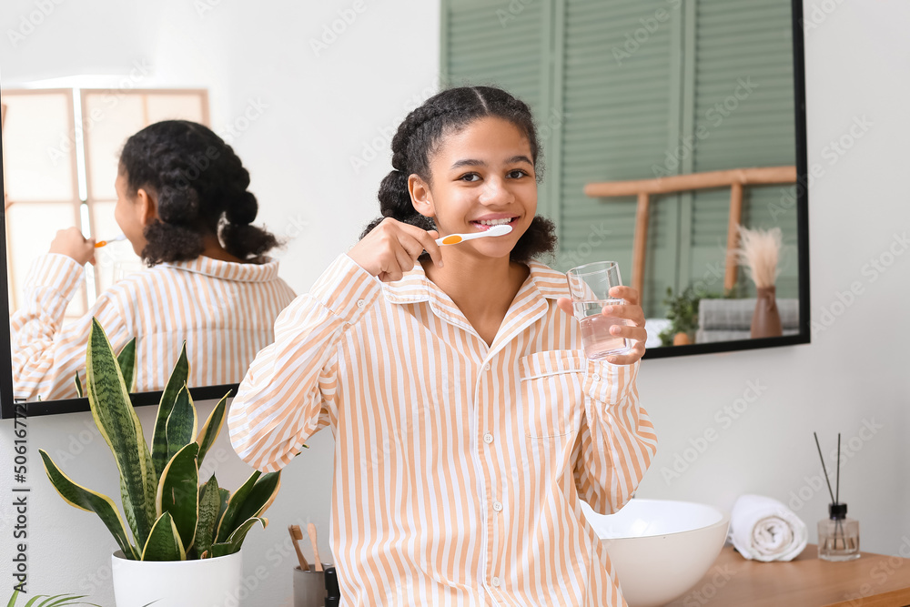 African-American teenage girl with glass of water brushing teeth in bathroom