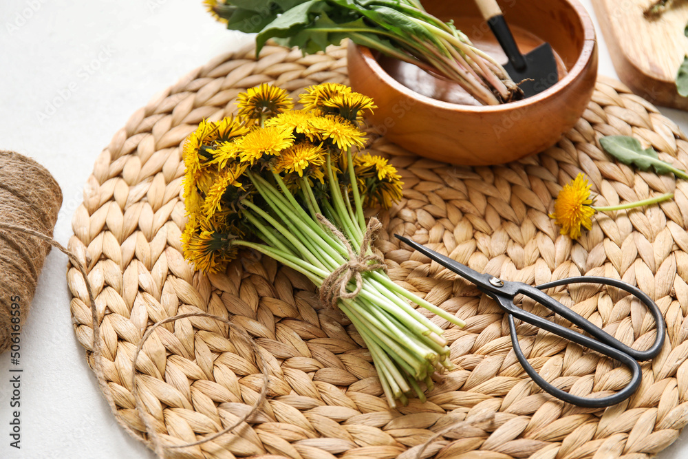 Wicker mat with dandelions, bowl, shovel and scissors on white background, closeup