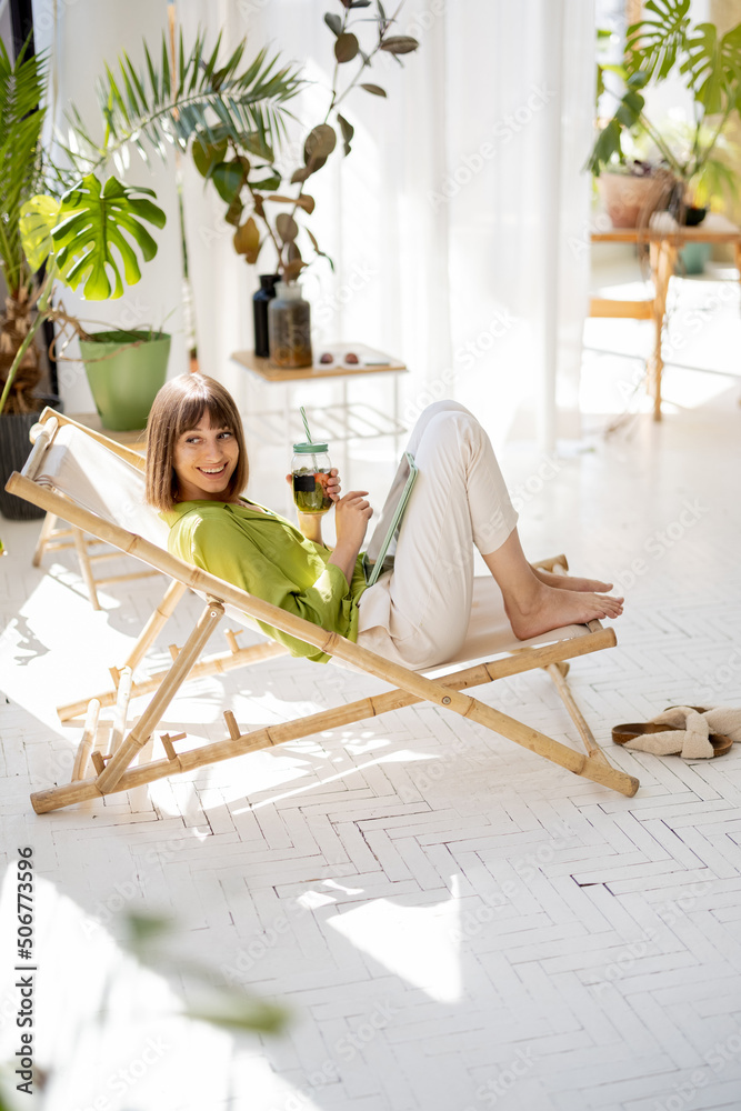 Woman sits relaxed with a drink on sunbed in sunny living room with green plants on background. Leis