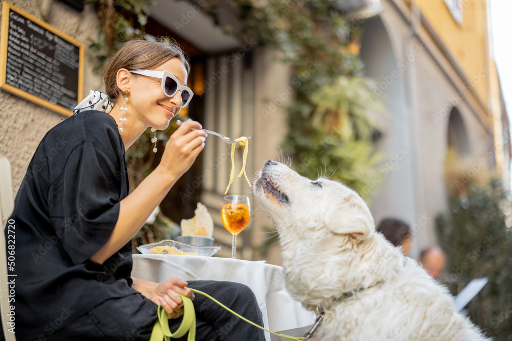 Woman eating pasta with her cute white dog at the restaurant outdoors. Concept of friendship with pe