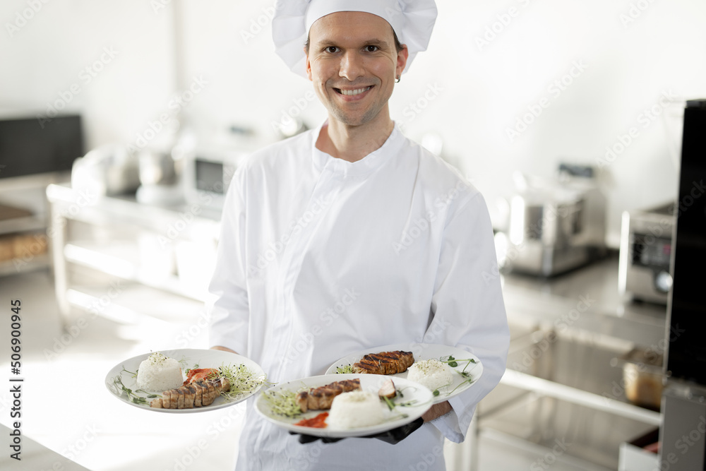 Portrait of young handsome smiling chef in uniform standing with ready meals in the kitchen. Happy c