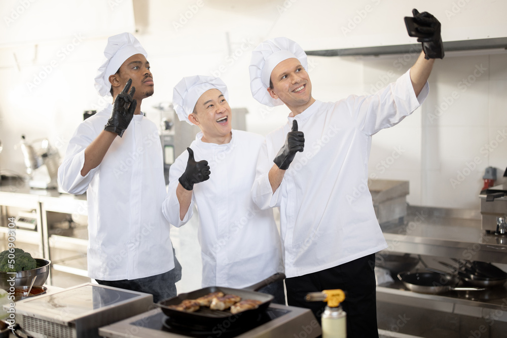 Happy multiracial team of cooks take selfie photo on phone while cooking together in the kitchen. Ch