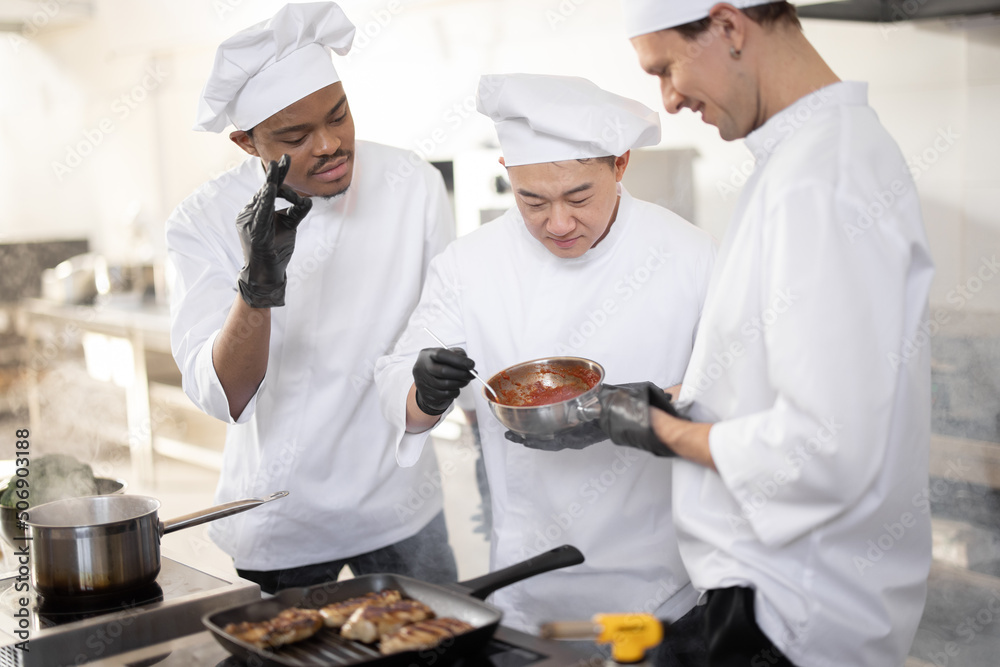 Three chef cooks with different ethnicities tasting sauce with a spoon while cooking in the kitchen.