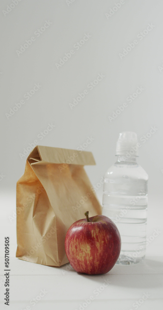 Vertical image of packed lunch in paper bag with apple and water on blue background