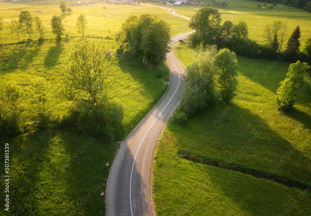 夏季日落时，绿色草地和山丘上的道路鸟瞰图。乡村道路的无人机俯视图