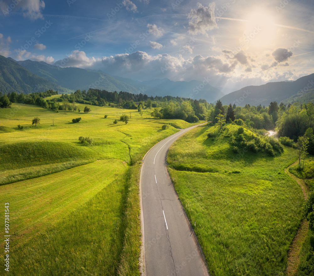 Aerial view of road in green meadows and hils at sunset in summer. Top view from drone of rural road