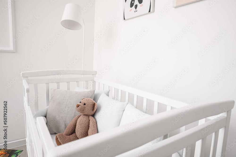 Baby crib with pillows and teddy bear near light wall