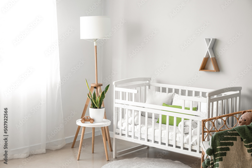 Interior of light nursery with baby crib, lamp, table and armchair