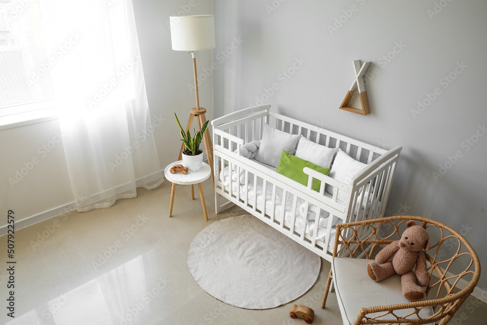 Interior of light nursery with baby crib, lamp, table and armchair