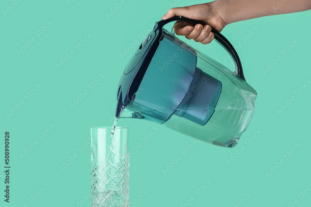 Woman pouring purified water into glass from filter jug on color background