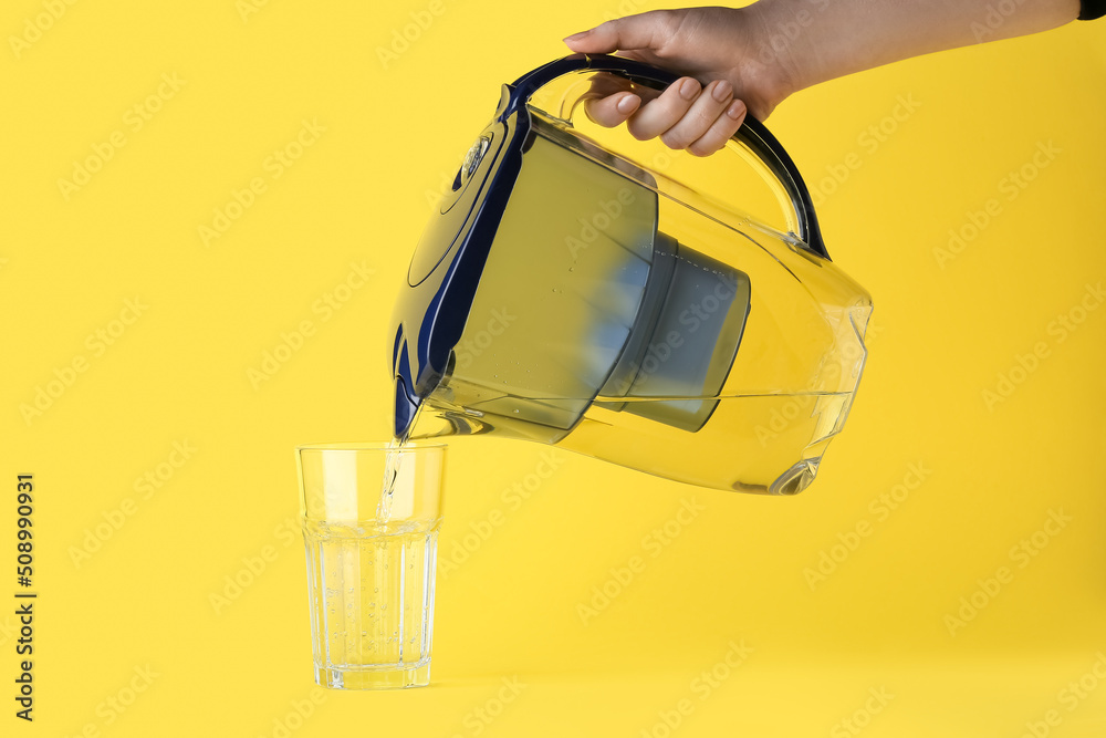 Woman pouring purified water into glass from filter jug on yellow background