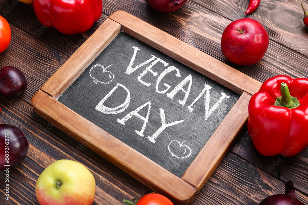 Different fresh products and board with text VEGAN DAY on wooden background, closeup