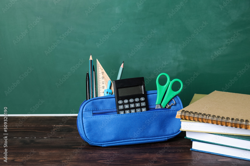 Pencil case with school stationery and books on table near green chalkboard