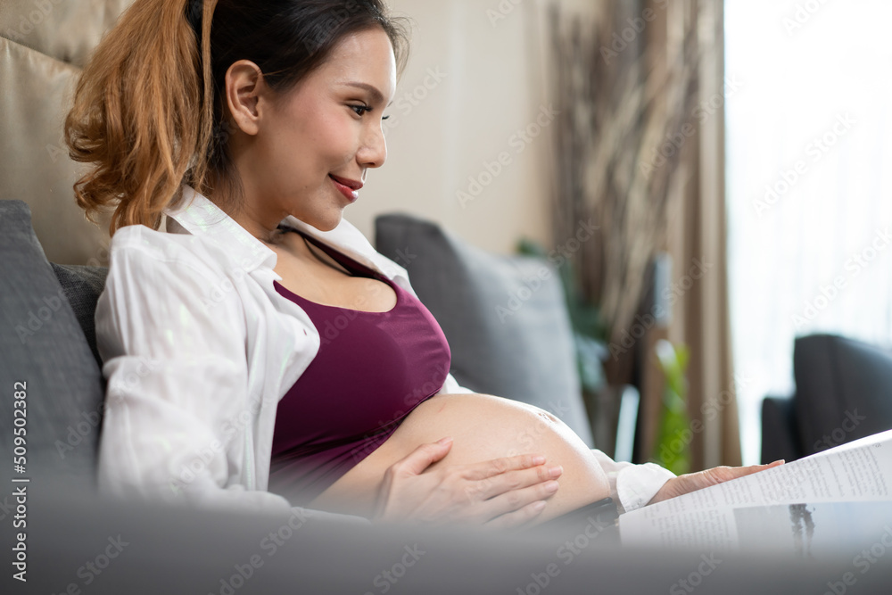 Asian young woman pregnant sitting and reading book with smile on sofa