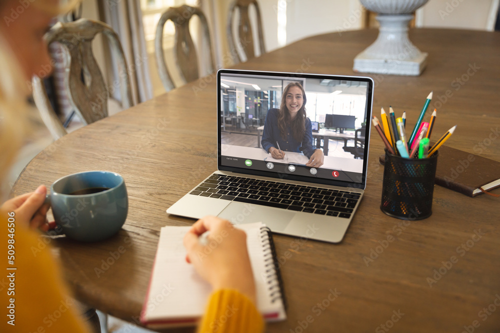 Happy caucasian businesswoman writing during video call with caucasian female colleague having drink