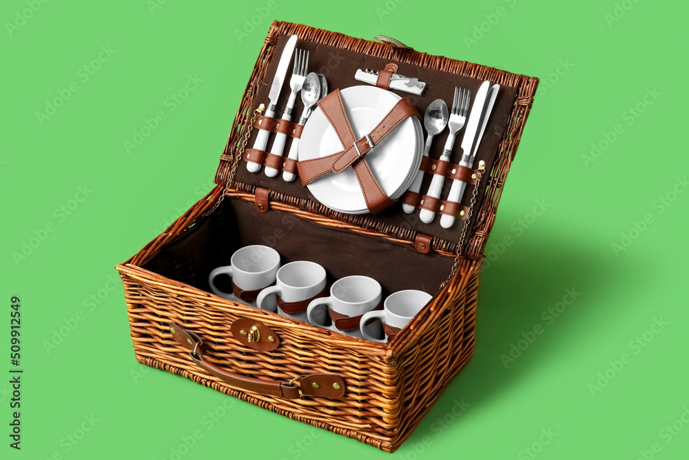 Wicker basket with tableware for picnic on green background