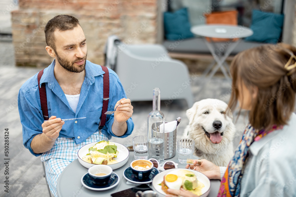 Stylish man and woman have a breakfast while sitting with dog at cafe outdoors