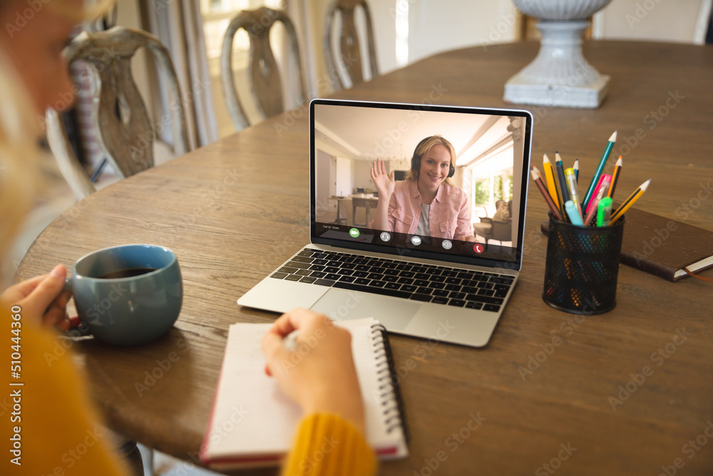 Caucasian businesswoman writing in notepad while video conferencing with teammate over laptop