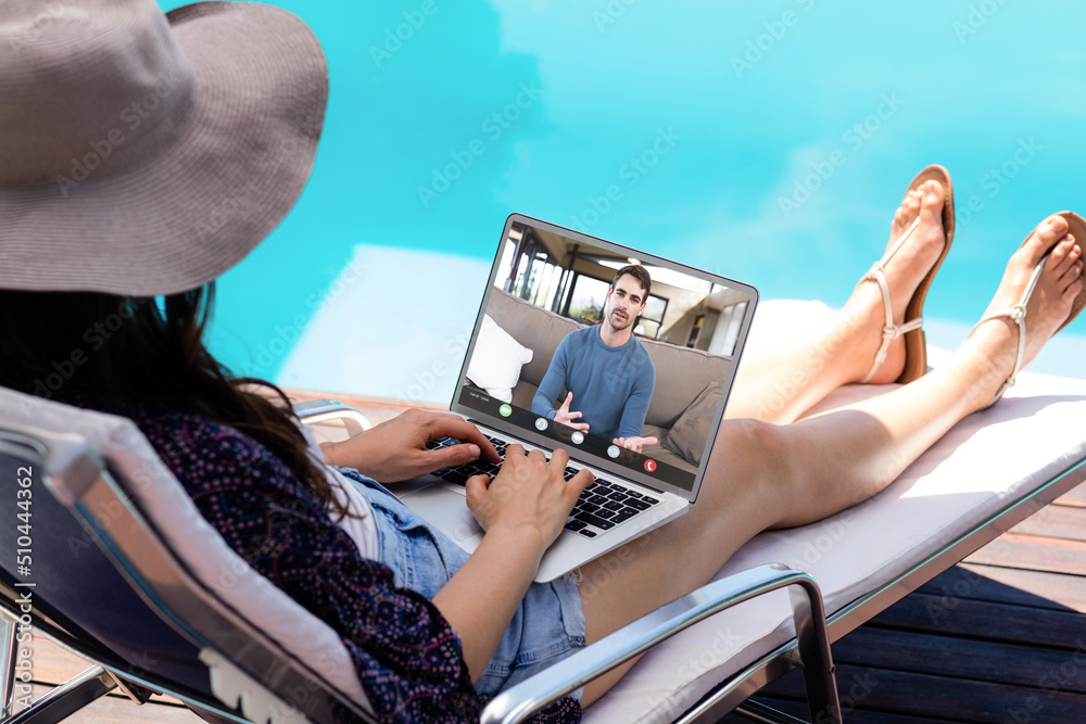 Caucasian young woman video calling male coworker through laptop on poolside during vacation