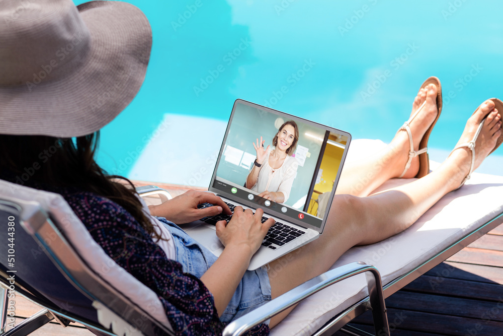 Caucasian businesswoman discussing with coworker over laptop while sitting at poolside on vacation