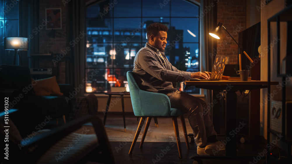 Young Handsome Black Man Working from Home on Laptop Computer in Stylish Loft Apartment in the Eveni