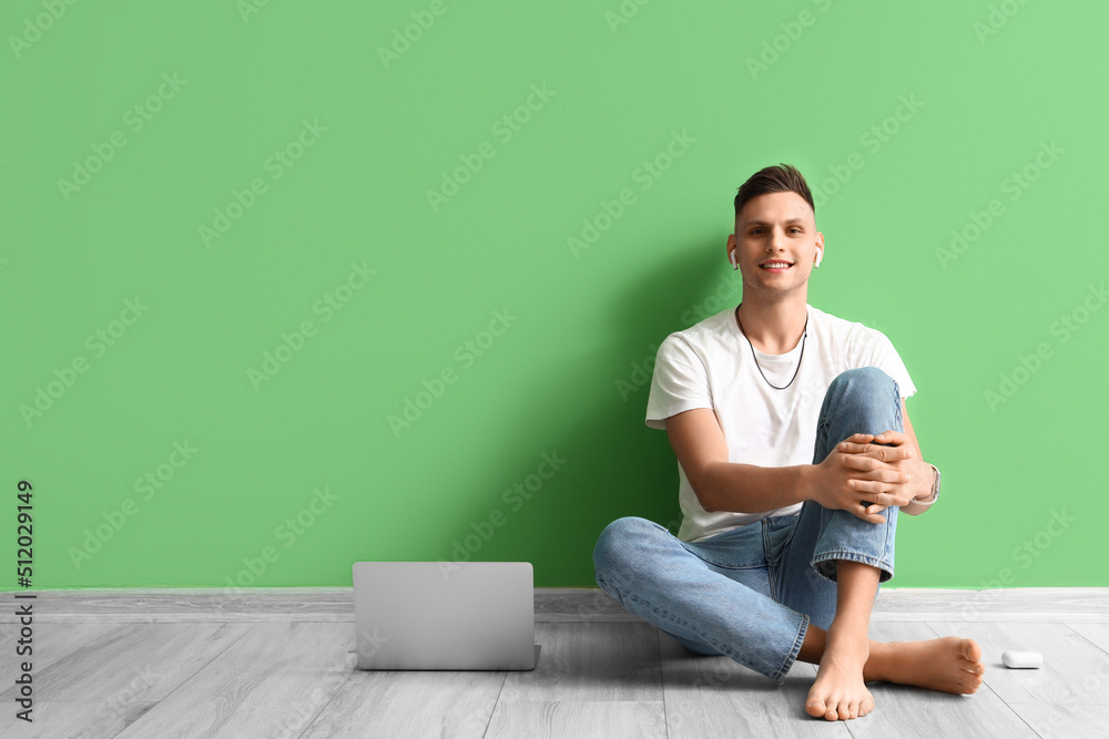 Young barefoot man with earphones and laptop near green wall