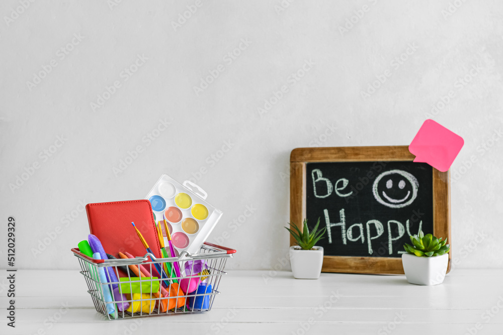 Shopping basket with different stationery and chalk board with phrase BE HAPPY on table near light w