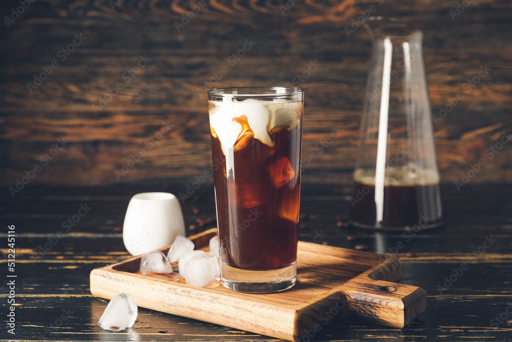 Board with glass of cold brew coffee and ice cubes on dark wooden table