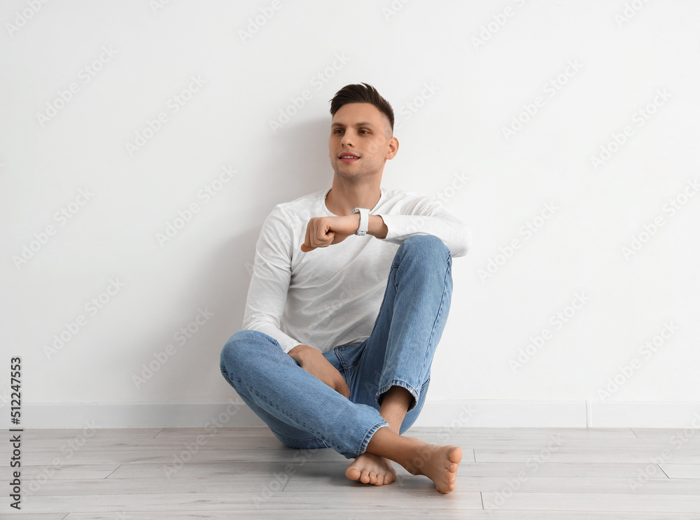 Young barefoot man with smartwatch sitting near light wall