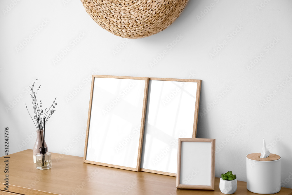 Blank frames, vase with flowers and tissue box on table near light wall