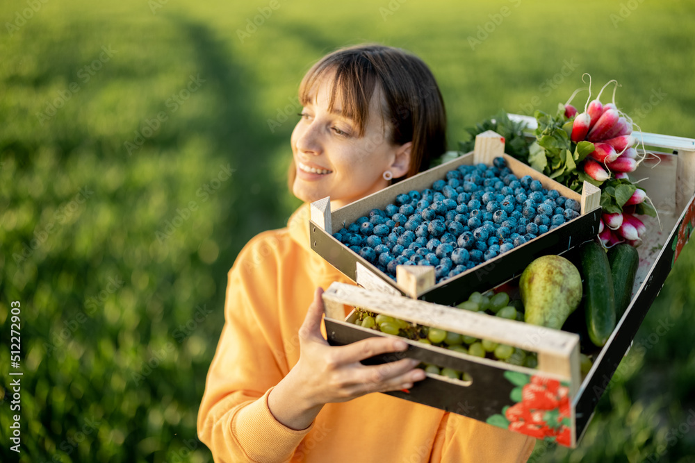 Cheerful woman carrying boxes with fresh juicy berries, fruits and vegetables on farmland. Concept o