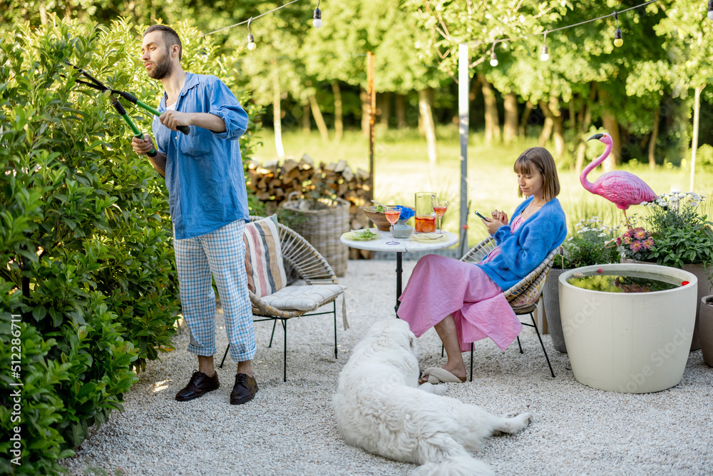 Young stylish family spend summer time at backyard of their country house on nature. Man pruning gre