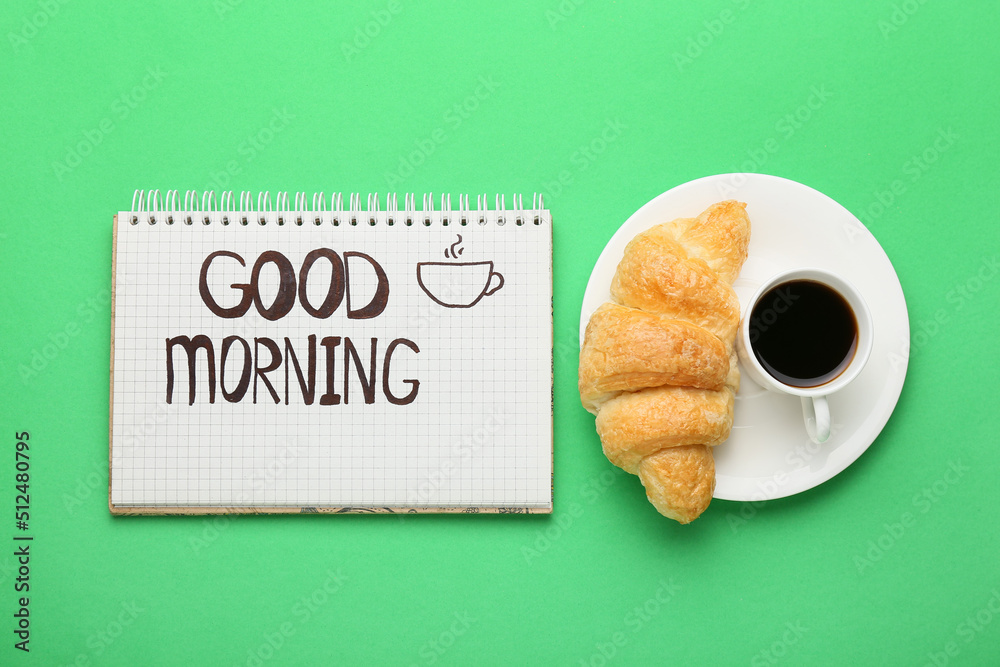 Notebook with text GOOD MORNING, cup of coffee and croissant on green background