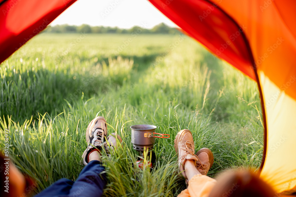 Couple sit in tent together and prepare some food on tourist burner, view on shoes and green field o