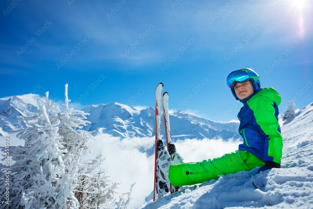 带着高山滑雪板的男孩坐在山峰和云朵上的雪地里