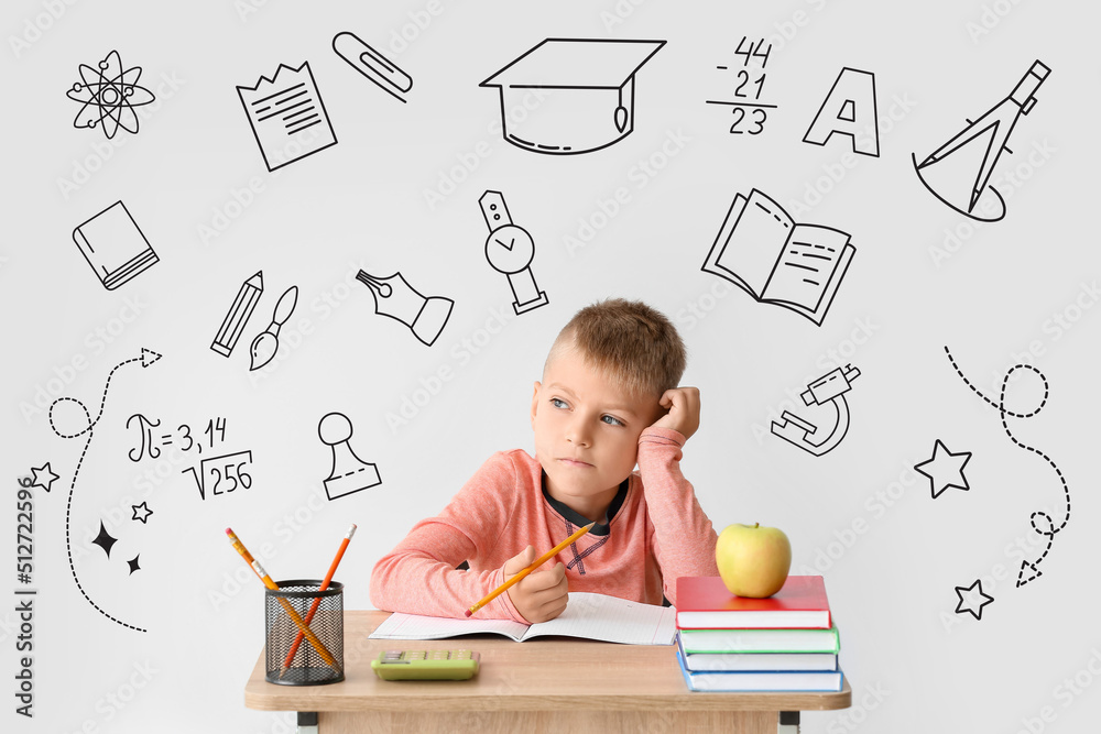 Thoughtful little schoolboy sitting at desk against light background