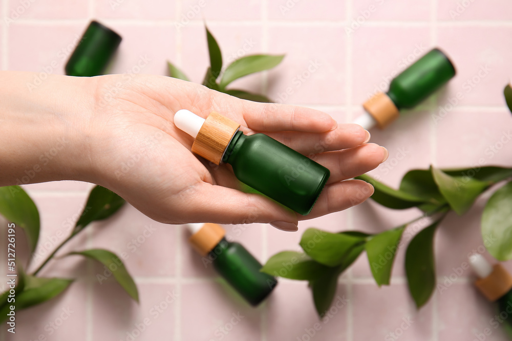 Female hand with bottle of natural serum and plant branches on color tile, closeup