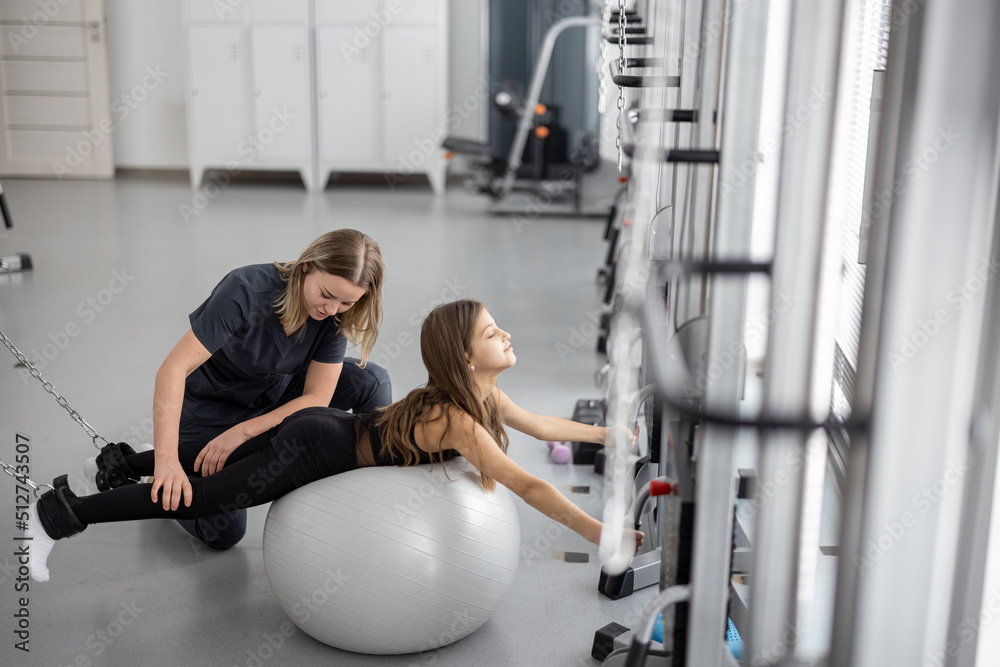 Rehabilitation specialist helping little girl to do exercises with fitness ball and decompression si