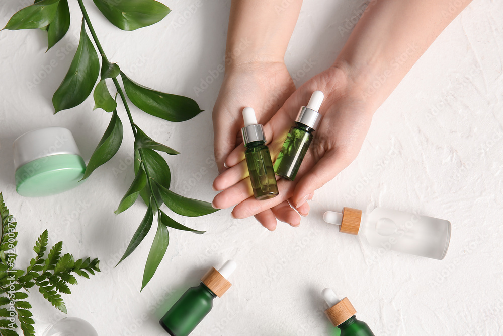 Female hands with bottles of natural serum, cosmetics and plant branch on light background