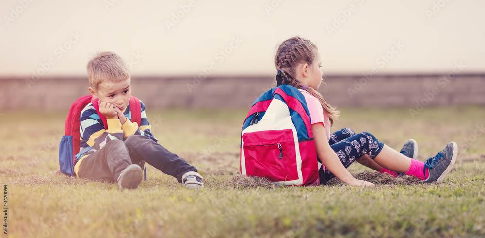 A boy and a girl quarreling and are sitting on the grass in the schoolyard back to back