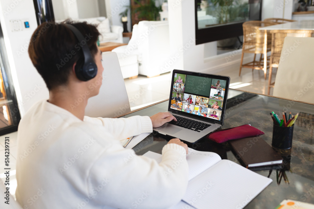 Asian teenage boy wearing headphones while learning online over laptop at home