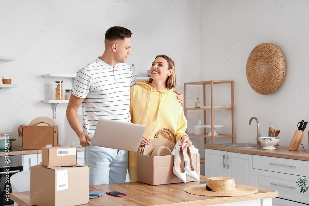 Young couple with laptop unpacking box with new clothes in kitchen. Online shopping