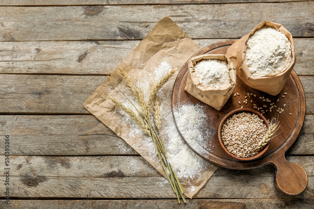 Wheat ears, bowl, paper bags with flour, board and parchment paper on wooden background