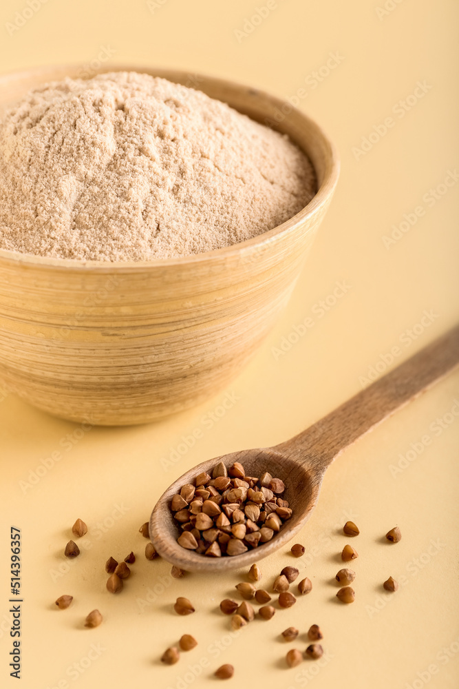 Bowl of flour and spoon with buckwheat grains on beige background