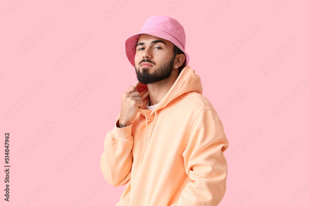 Fashionable bearded man wearing hoodie and hat on pink background